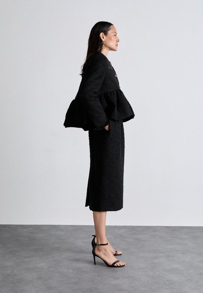 Black peplum jacket with textured fabric and three gold buttons, paired with a fitted black midi skirt and strappy black high-heeled sandals.