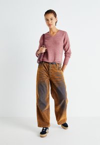 Rosa gerippter Pullover mit V-Ausschnitt. Braune weite Cordhosen mit kontrastierenden Mustern und Knöpfen. Schwarze Sneakers und eine kleine Tasche.