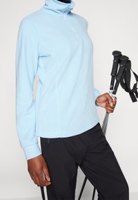 Pólvora de forro polar de color azul claro con cuello de media cremallera, mangas largas y costuras planas. Llevado con pantalones negros, sosteniendo bastones de senderismo.
