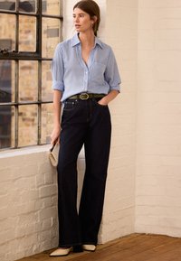 Mėlynos ir baltos dryžuotos marškiniai su užsegamais mygtukais ir sukištais rankovėmis, derinami su tamsiai mėlynais plačiomis kojomis džinsais. Papuošti žalia diržu ir smėlio spalvos delninuke.