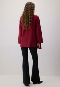 Burgundia pikkade varrukatega pluus, mis on lõdva lõikega, kombineeritud mustade lehvikpükstega, nähtud tagantpoolt. Sile kangas minimaalse disainiga.