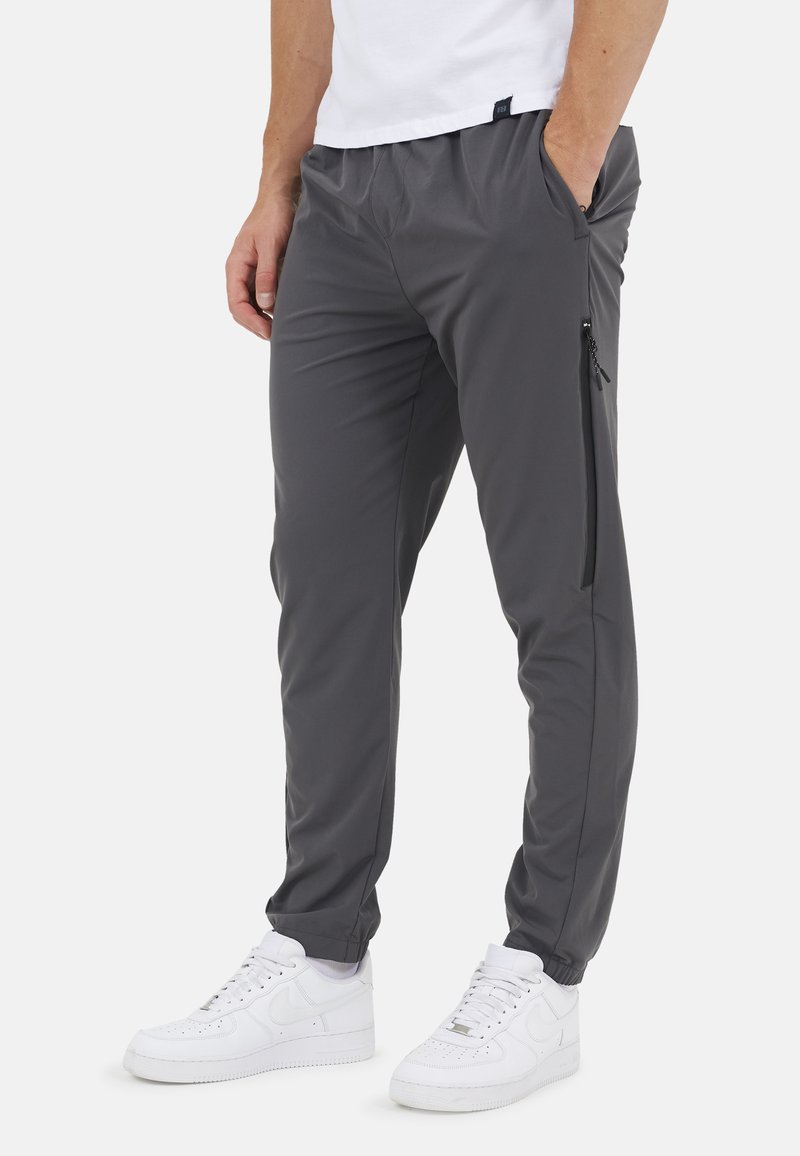 Uomo con pantaloni casual grigio scuro con tasca laterale con cerniera e sneakers bianche, in piedi con una mano in tasca.