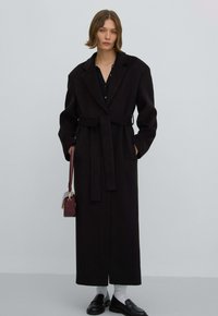 Zwarte wollen jas met een lange, rechte silhouet, grote revers en een strikceintuur. Draagt het over een zwart topje, gecombineerd met loafers en een kleine handtas.