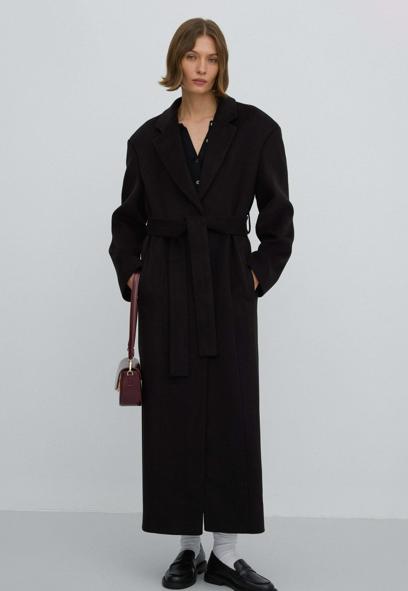 Zwarte wollen jas met een lange, rechte silhouet, grote revers en een strikceintuur. Draagt het over een zwart topje, gecombineerd met loafers en een kleine handtas.