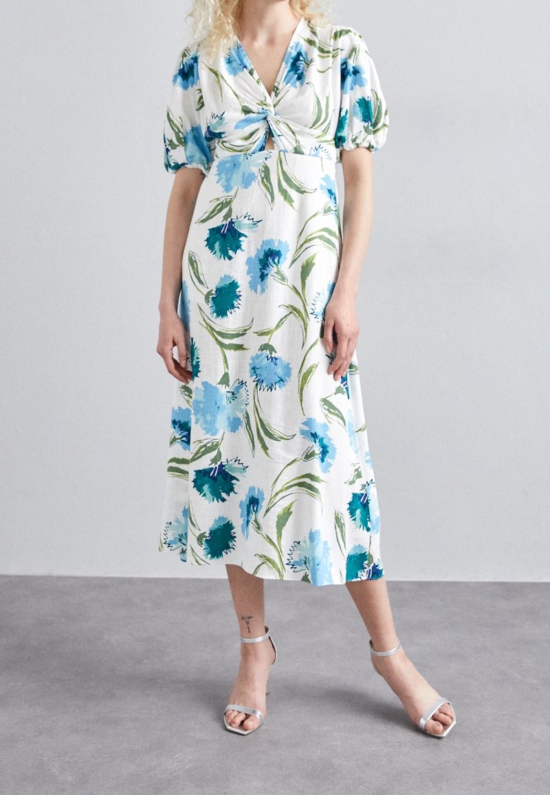 Femme portant une robe midi blanche à imprimé floral bleu et vert, manches bouffantes, nœud devant, et talons argentés à bride à la cheville, debout sur un sol gris.