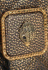 Zwart leer met gouden vlekken en een decoratieve metalen gesp in de vorm van een gestileerd blad, geaccentueerd met strass-steentjes.