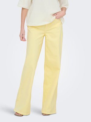 Femme portant un pantalon large jaune pâle et un chemisier crème, debout avec une main dans la poche devant un arrière-plan uni.