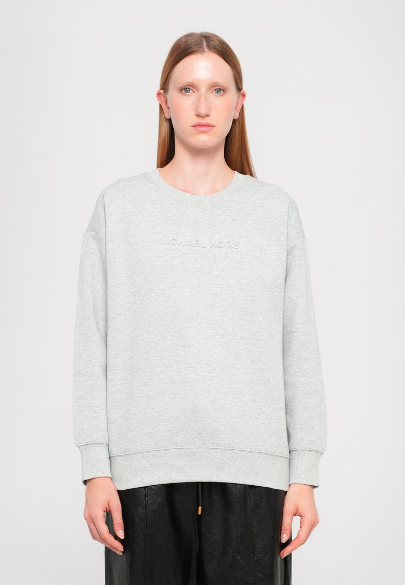 Sudadera gris hecha de un tejido suave, con cuello redondo y hombros caídos. El nombre de la marca "MICHAEL KORS" está grabado en la parte frontal.