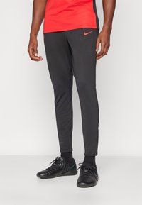 Nike treniruotės kelnės juodos spalvos, su liesu siluetu, lygiu audiniu, šoninėmis skeltukų detalėmis ir raudonuoju logotipo akcentu šalia klubo.