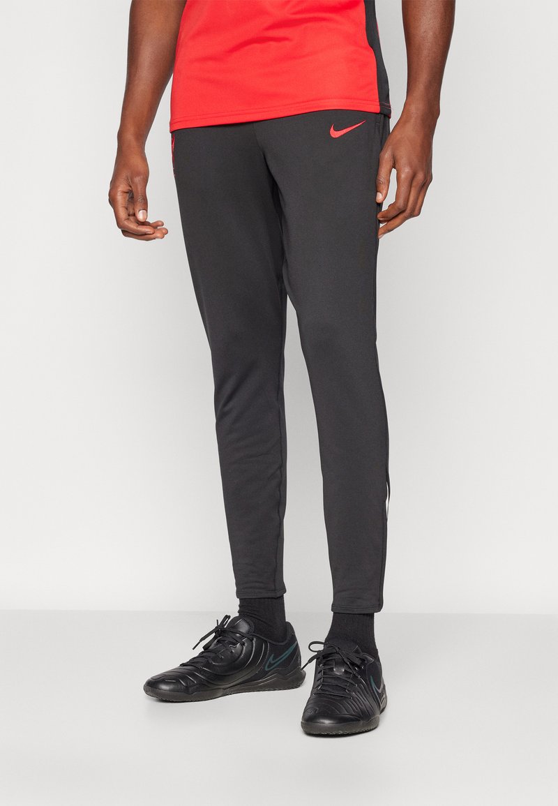 Nike treniruotės kelnės juodos spalvos, su liesu siluetu, lygiu audiniu, šoninėmis skeltukų detalėmis ir raudonuoju logotipo akcentu šalia klubo.