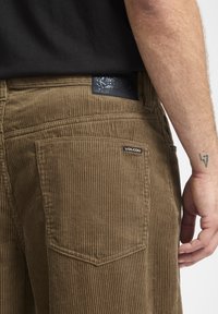 Brune kordfløjlsbukser med lodret ribbning, der har et Volcom-logo på baglommen og et mønstret label i taljen.