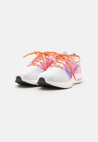 Baskets de sport blanches avec des lacets orange, des accents rayés roses et bleus, et des semelles noires, présentées côte à côte sur un fond blanc.