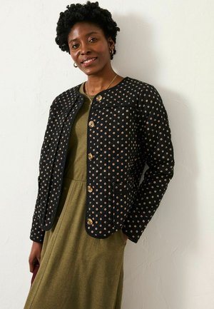 Schwarze gesteppte Jacke mit goldenen, runden Mustern und Knopfverschlüssen, kombiniert mit einem grünen Kleid. Einfacher Kragen und seitliche Taschen.