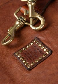 Accessorio in pelle con una superficie marrone scuro testurizzata, dotato di ganci in metallo e di un'etichetta rettangolare con borchie dorate e testo impresso.