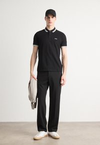 Schwarzes Poloshirt mit kontrastierendem Kragen und Besatz, kombiniert mit schwarzen Hosen und weißen Sneakers. Grauer Strickpullover über dem Armgetragen.