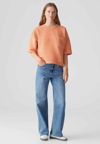 Top oversize a maniche corte con texture a coste di colore corallo, dotato di taschino, abbinato a jeans a gamba larga di colore blu chiaro e scarpe slip-on grigie.