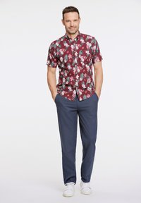 Camicia a maniche corte bordeaux con motivo floreale e foglie verdi, abbinata a pantaloni blu navy e Sneakers bianche. Focalizzarsi su motivi vividi e vestibilità rilassata.