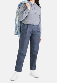 Grå hoodie med snörning, kombinerad med höga marinblå byxor och vita sneakers. En blå rutiga skjorta hänger över ena axeln.