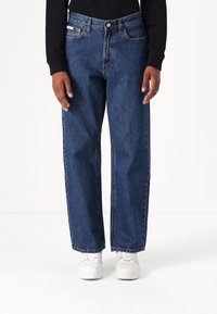 Jeans de mezclilla azul con un corte holgado, cinco bolsillos y detalles de costura visibles. Llevados con zapatillas blancas y una blusa de manga larga negra.