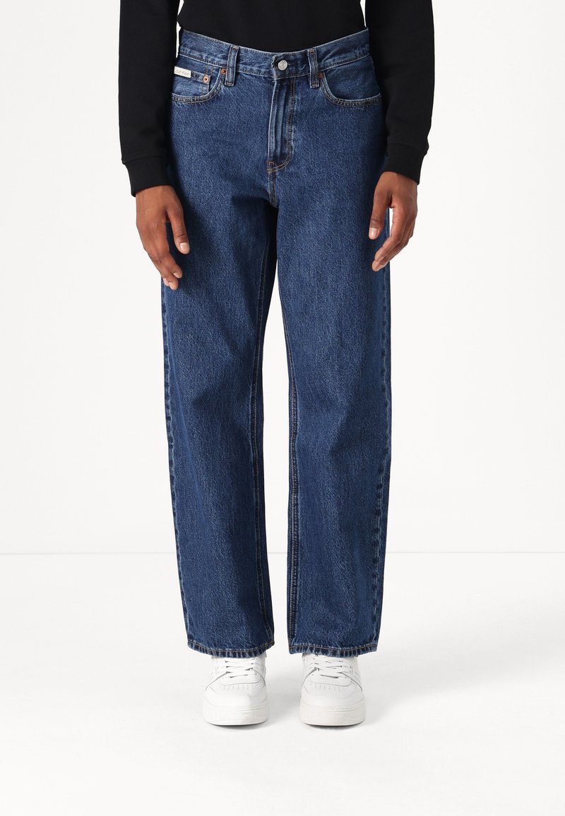 Jeans de mezclilla azul con un corte holgado, cinco bolsillos y detalles de costura visibles. Llevados con zapatillas blancas y una blusa de manga larga negra.