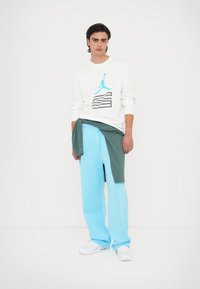 Camicia bianca a maniche lunghe con un design grafico blu, abbinata a pantaloni della tuta azzurri e una felpa verde scuro legata in vita.