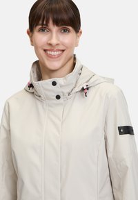 Veste imperméable beige clair avec un col haut, capuche ajustable, accents noirs et cordons de serrage roses contrastants. Texture lisse, design élégant.
