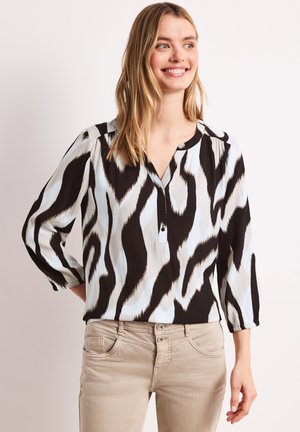 Uśmiechnięta kobieta w czarno-białej bluzce z abstrakcyjnym wzorem z rękawami do łokcia i beżowych spodniach, stojąca na jednolitym tle.