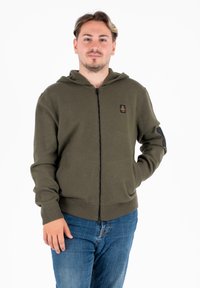 Felpa con zip di colore verde oliva, con cappuccio, polsini e orlo a costine; presenta un piccolo logo nero sul petto e toppe nere sui gomiti.