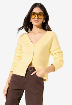 Yellow wool cardigan with a v-neck, long sleeves, and button closure, paired with brown pants and accessorized with sunglasses and earrings.