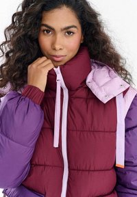 Kobieta o kręconych ciemnych włosach, ubrana w fioletowo-bordową pikowaną kurtkę, trzymająca suwak blisko podbródka, patrząca prosto przed siebie.