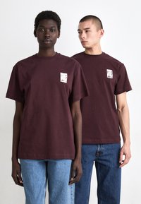 Burgunderfargede bomulls T-skjorter med korte ermer, med et lite grafisk trykk på brystet. Modellene parer med blå denimjeans.