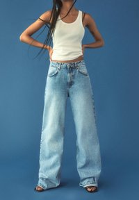 Personne portant un débardeur blanc, un jean taille haute à jambes larges et des sandales noires à bouts ouverts sur un fond bleu, mains sur les hanches.