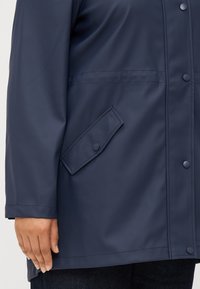 Close-up of a person's hand resting on the hem of a navy waterproof jacket, featuring a buttoned flap pocket and snap closures.