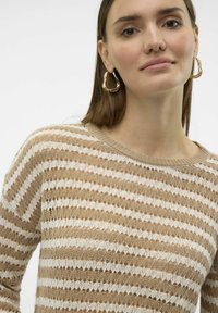 Pull en tricot rayé beige et blanc avec une texture ajourée. Encolure ronde et coupe ajustée. Modèle portant des boucles d'oreilles en or.