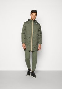 Green puffer coat with a hood, featuring a yellow and red stripe down the front. Worn over matching green pants and black sneakers.