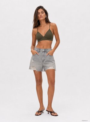 Donna che indossa un bralette verde oliva, pantaloncini di jeans grigio chiaro usurati e sandali neri con tacco, in piedi con le mani nelle tasche.