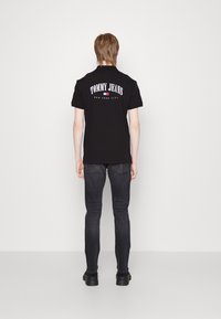 Svart bomullspolo med märkeslogotypen "TOMMY JEANS" på ryggen. Kombinerad med slim-fit mörka jeans och svarta skor. Enkel, avslappnad design.
