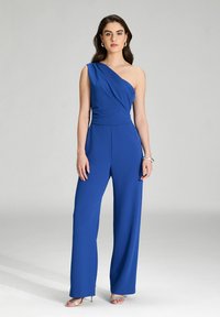 Combinaison une épaule bleue en tissu lisse, avec un corsage drapé, des pantalons à jambes larges et de subtils plis pour une texture.