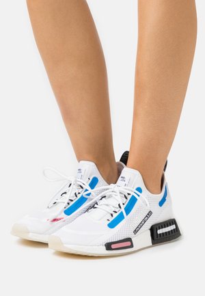 Baskets de sport blanches avec des accents bleu et noir, partie supérieure en maille, semelle texturée et détail contrastant rose sur le côté.