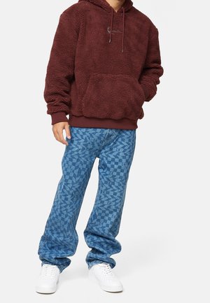 Persona con indossata una felpa con cappuccio in sherpa color granata, jeans a zampa larghi con motivo blu e sneakers bianche, in piedi con una mano in tasca.