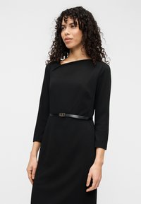 Robe noire à manches longues avec un col bateau, dotée d'une taille cintrée soulignée par une fine ceinture noire avec un détail de boucle métallique.