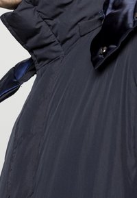 Giacca imbottita blu navy con collo alto, caratterizzata da un dettaglio in raso e chiusura con bottoni. Texture liscia con design leggero.