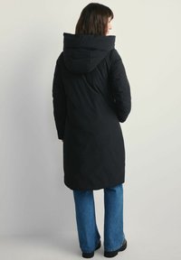 Kobieta ubrana w długi czarny zimowy płaszcz z kapturem, w niebieskie dżinsy i czarne buty, stoi plecami do obiektywu na jednolitym tle.