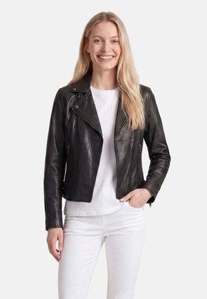 Lächelnde Frau mit langen blonden Haaren, die eine schwarze Lederjacke, ein weißes T-Shirt und weiße Hosen trägt, steht vor einem schlichten Hintergrund.
