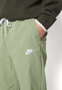 Osoba oblečená v olivově zelených kalhotách Nike s bílým logem, s rukou v kapse, v kombinaci s tmavě zeleným pleteným svetrem s bílými manžetami.