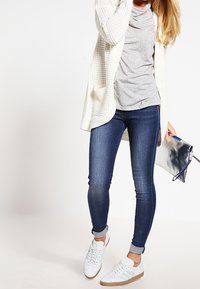 Frau in weißer Strickjacke, grauem Shirt, blauen Skinny-Jeans und weißen Turnschuhen, die in der rechten Hand eine silberne Clutch hält.