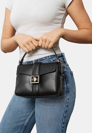 Donna con top bianco e jeans blu tiene per la maniglia una piccola borsa nera in pelle con chiusura dorata.