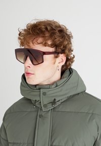 Røde oversized solbriller med gradientlinser, brukt med en grønn polstret jakke med høy krage og knappedetaljer.