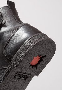 Bottine en cuir noir avec surface texturée, semelle en caoutchouc et accent rouge à travers une découpe. Présente des coutures et une boucle à l'arrière.