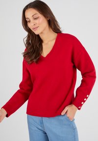 Maglione rosso lavorato a maglia con scollo a V, polsini a costine e bottoni decorativi sulle maniche. Il tessuto ha una consistenza morbida. Abbinato a jeans azzurri chiari.